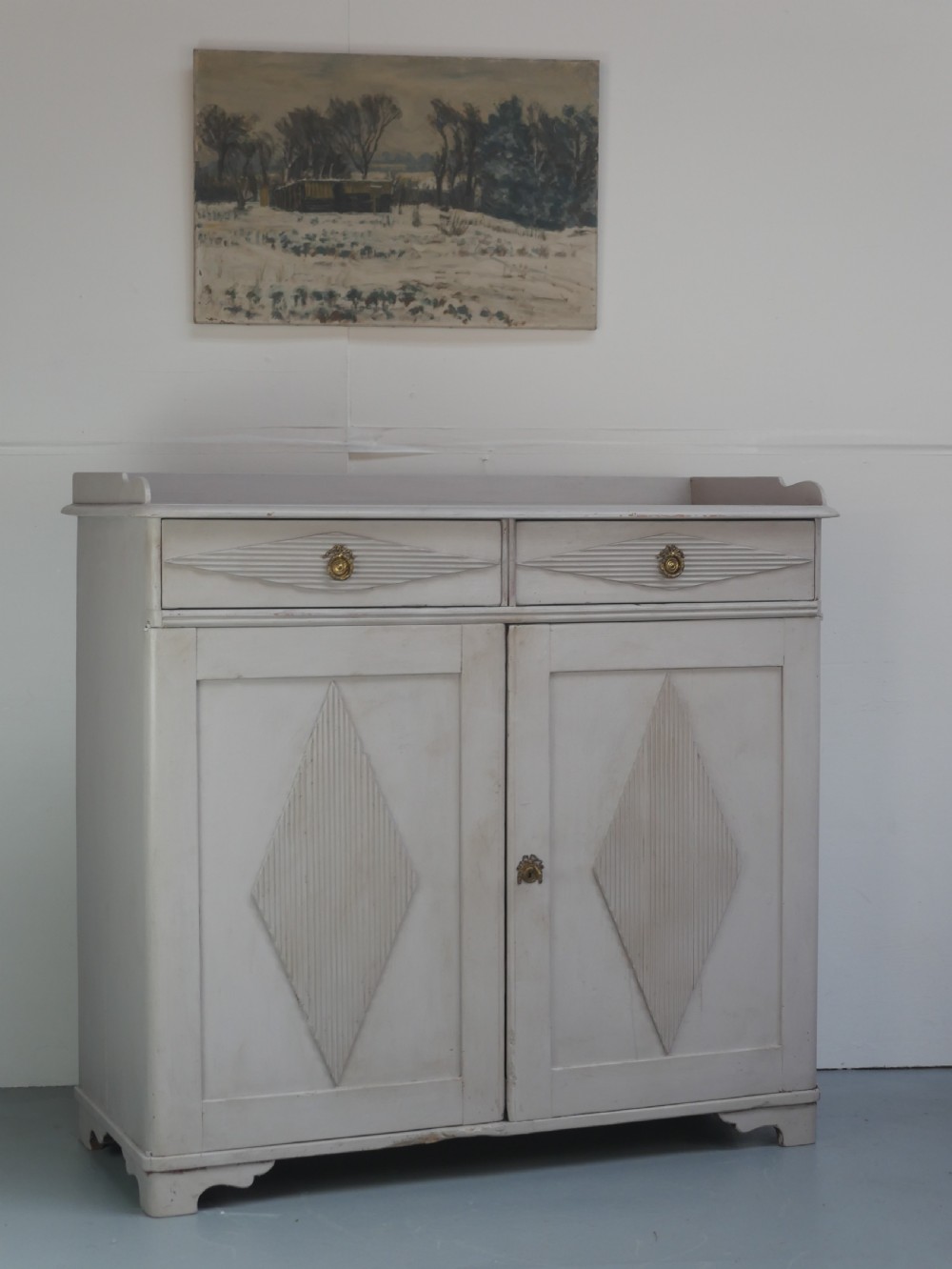 A pale grey painted Scandinavian cupboard featuring two carved drawers above two diamond-panelled doors with brass hardware.