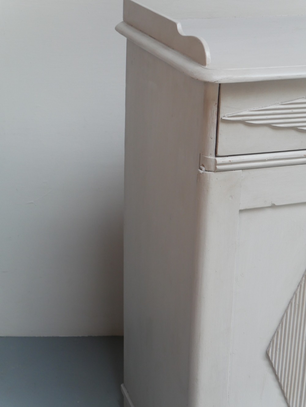 A pale grey painted Scandinavian cupboard featuring two carved drawers above two diamond-panelled doors with brass hardware. View X