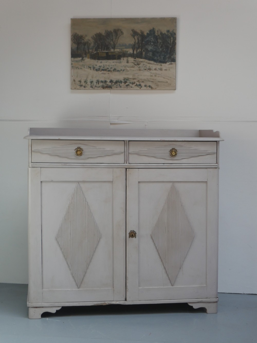 A pale grey painted Scandinavian cupboard featuring two carved drawers above two diamond-panelled doors with brass hardware. View XV