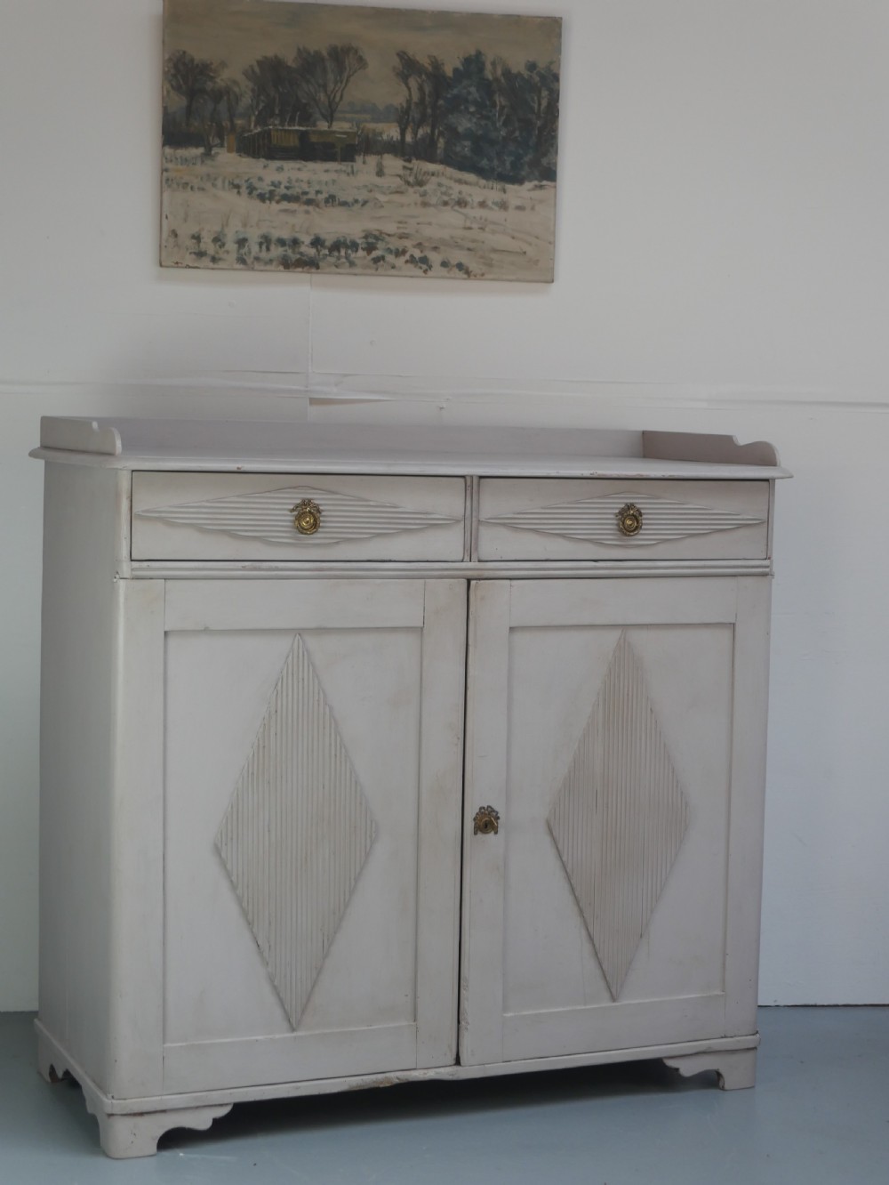 A pale grey painted Scandinavian cupboard featuring two carved drawers above two diamond-panelled doors with brass hardware. View XVI