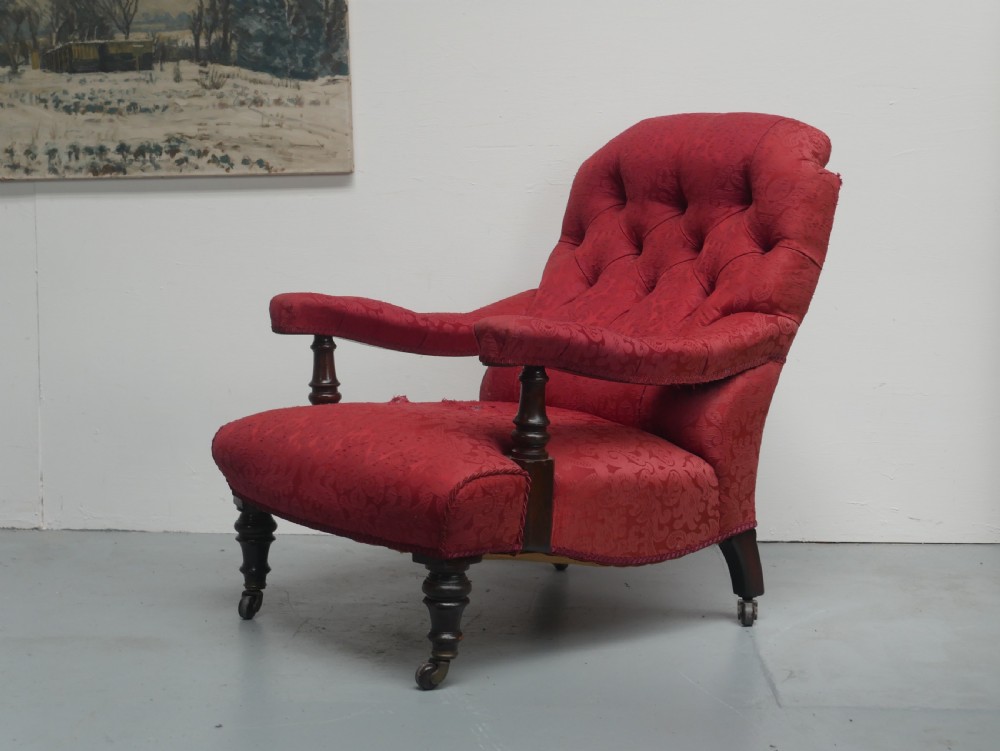 An antique Victorian Howard style armchair upholstered in red patterned damask fabric featuring a button-tufted backrest, low seat, and turned front wooden legs on brass castors.