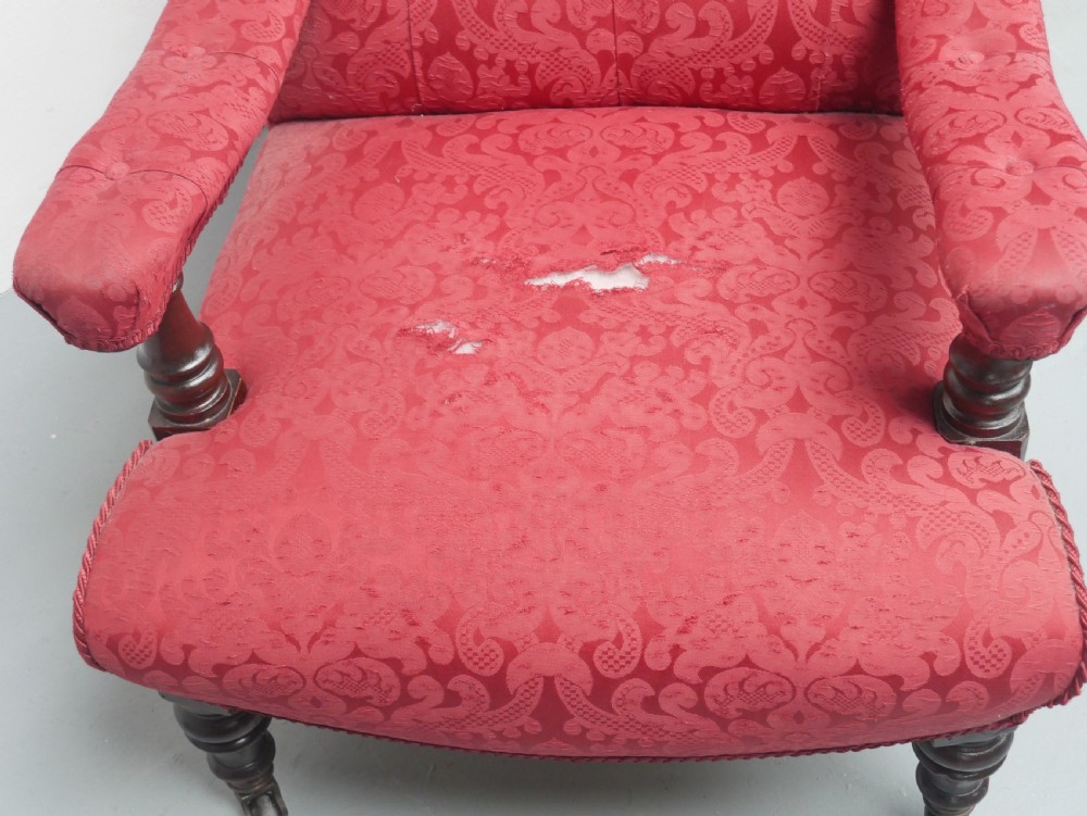 An antique Victorian Howard style armchair upholstered in red patterned damask fabric featuring a button-tufted backrest, low seat, and turned front wooden legs on brass castors. View II
