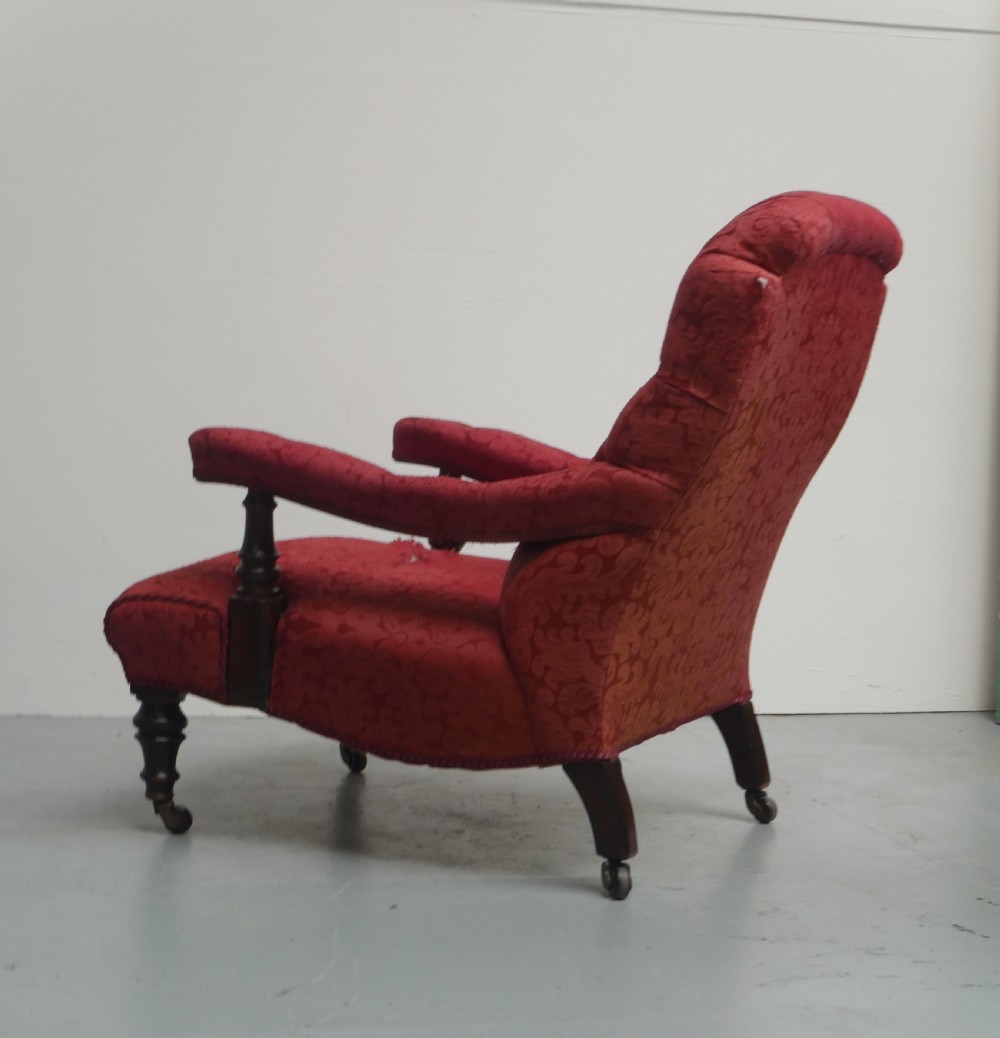 An antique Victorian Howard style armchair upholstered in red patterned damask fabric featuring a button-tufted backrest, low seat, and turned front wooden legs on brass castors. View IV