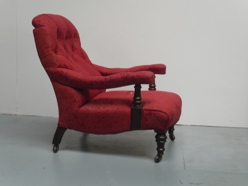 An antique Victorian Howard style armchair upholstered in red patterned damask fabric featuring a button-tufted backrest, low seat, and turned front wooden legs on brass castors. View V