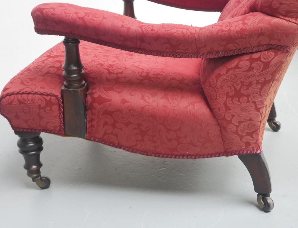 An antique Victorian Howard style armchair upholstered in red patterned damask fabric featuring a button-tufted backrest, low seat, and turned front wooden legs on brass castors. View VI