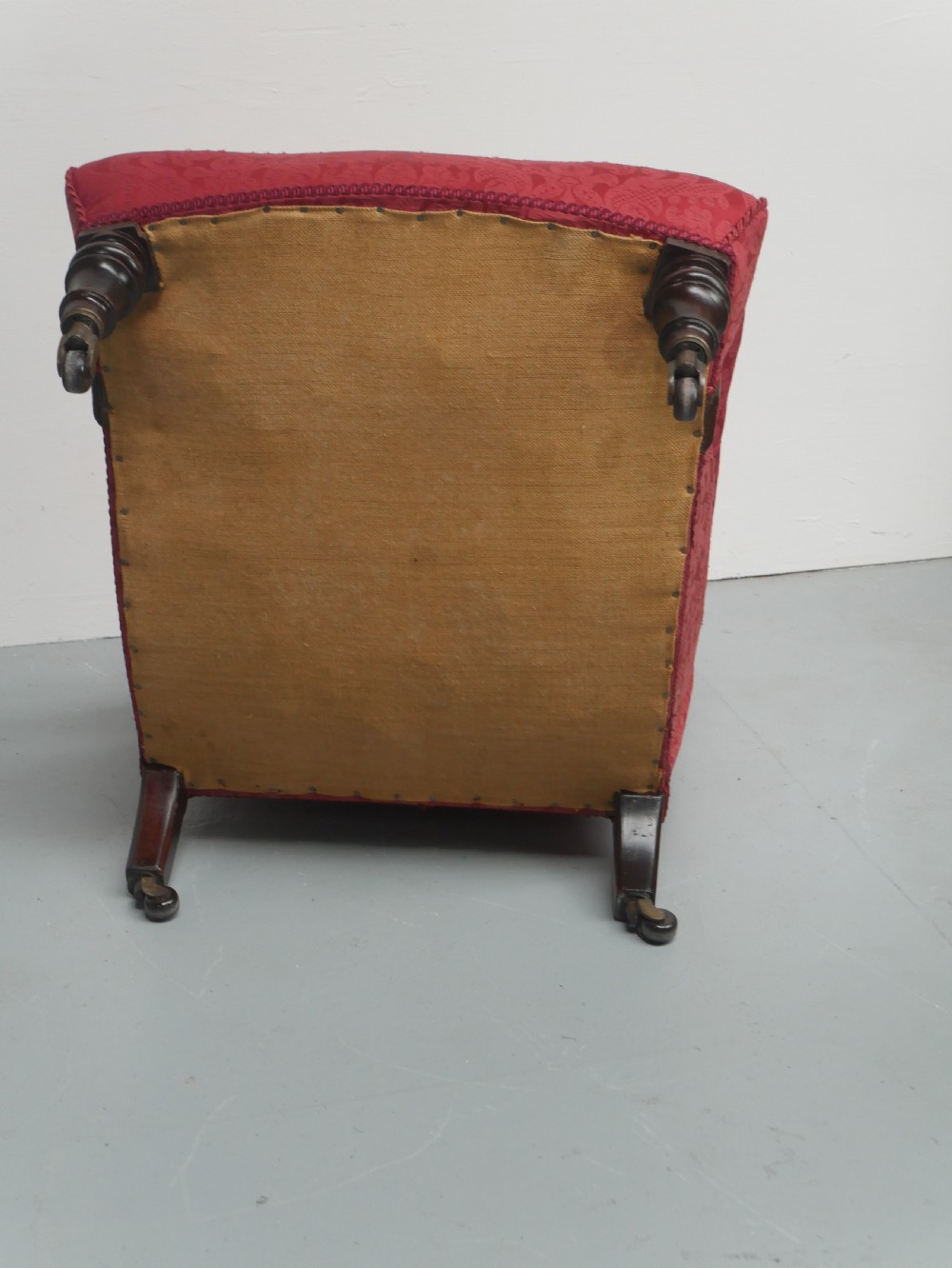 An antique Victorian Howard style armchair upholstered in red patterned damask fabric featuring a button-tufted backrest, low seat, and turned front wooden legs on brass castors. View VII