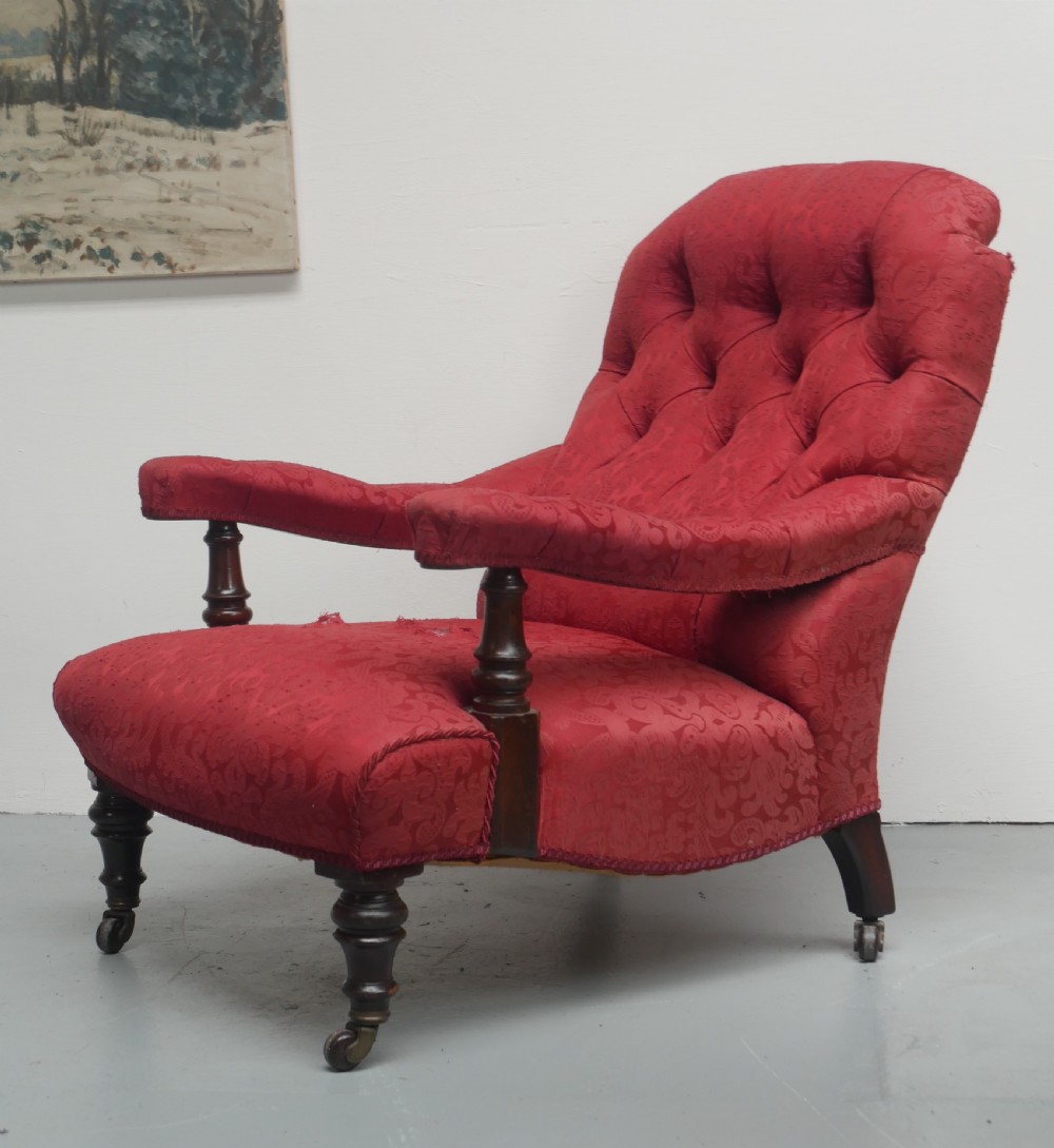 An antique Victorian Howard style armchair upholstered in red patterned damask fabric featuring a button-tufted backrest, low seat, and turned front wooden legs on brass castors. View XIII