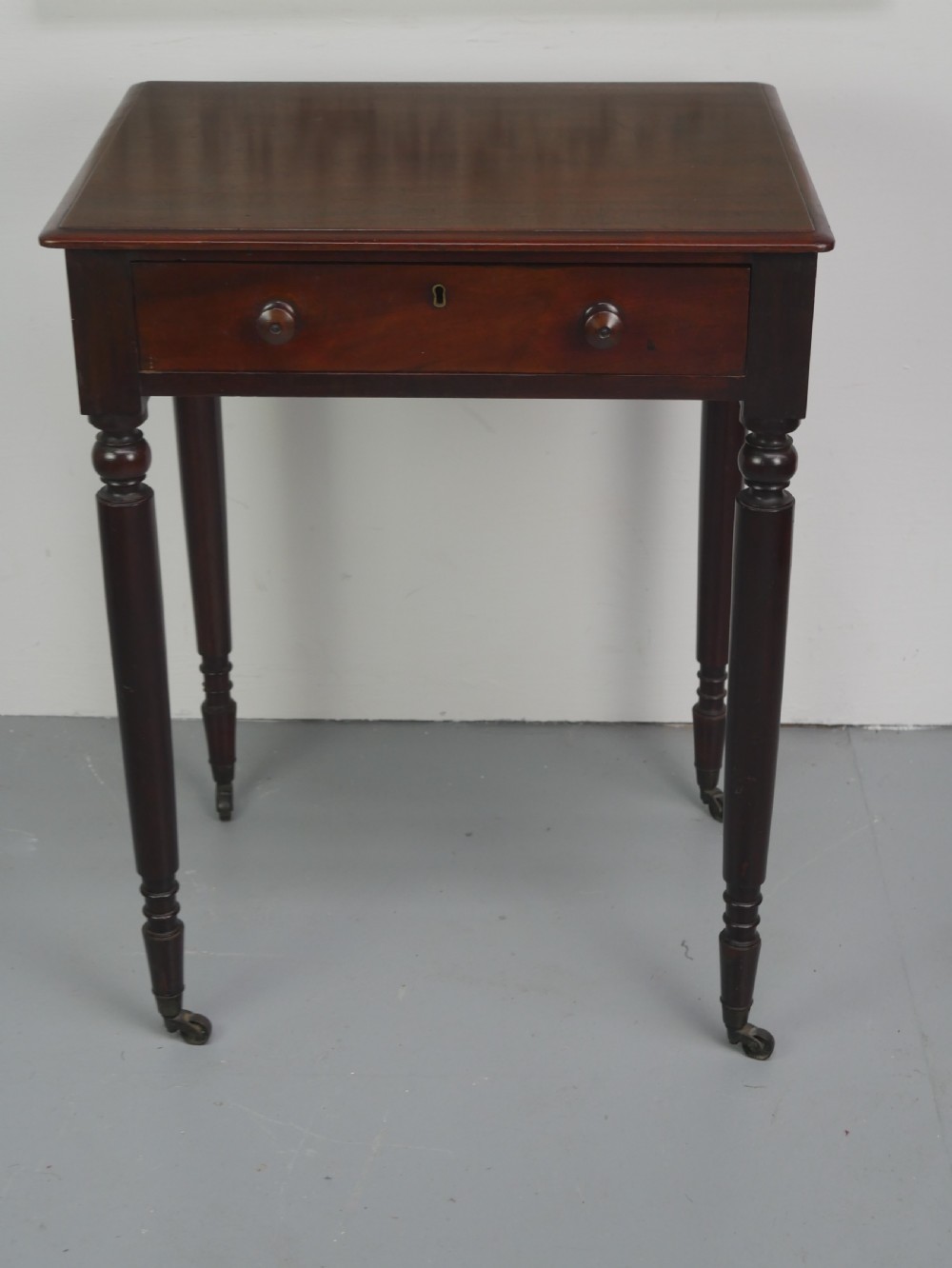 A dark mahogany Regency side table featuring a single drawer with turned knobs, a central brass keyhole, and turned legs terminating in castors. View II
