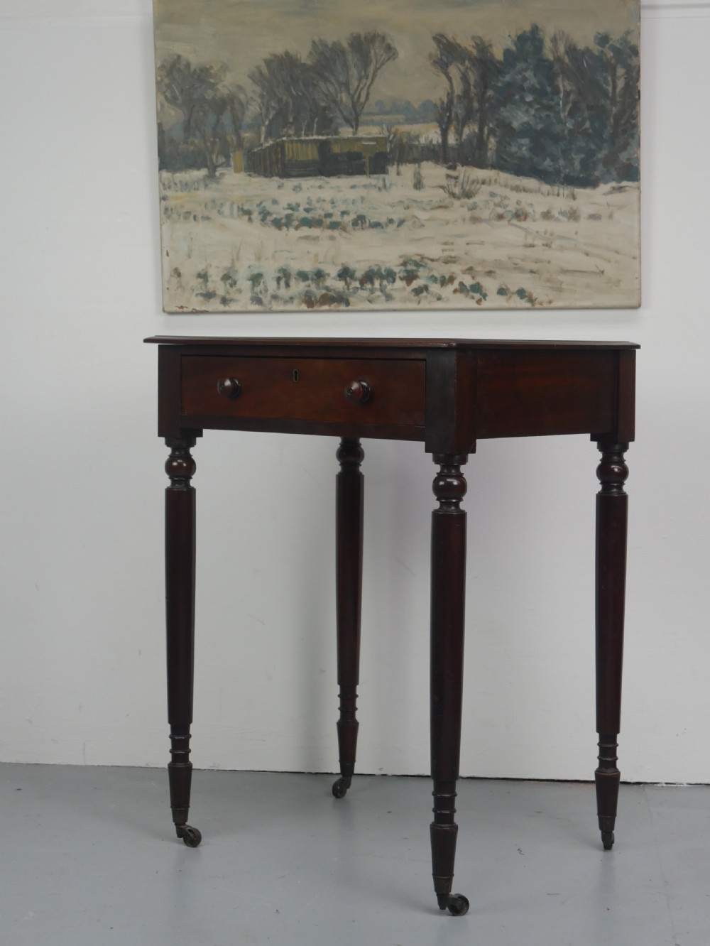 A dark mahogany Regency side table featuring a single drawer with turned knobs, a central brass keyhole, and turned legs terminating in castors. View III