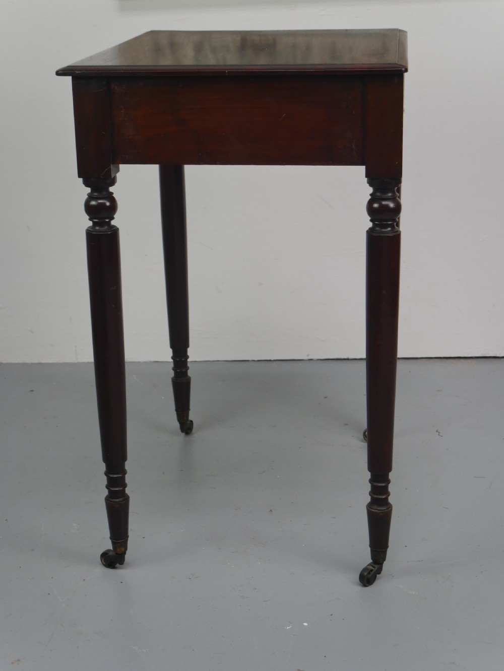 A dark mahogany Regency side table featuring a single drawer with turned knobs, a central brass keyhole, and turned legs terminating in castors. View VIII