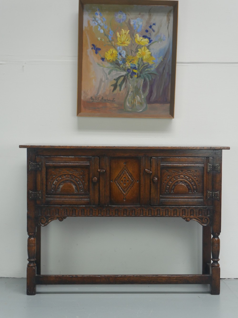A small dark oak carved sideboard featuring panelled cupboard doors with carved arch motifs, iron hinges, and turned legs with a stretcher base.