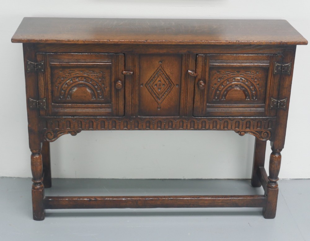 A small dark oak carved sideboard featuring panelled cupboard doors with carved arch motifs, iron hinges, and turned legs with a stretcher base. View II