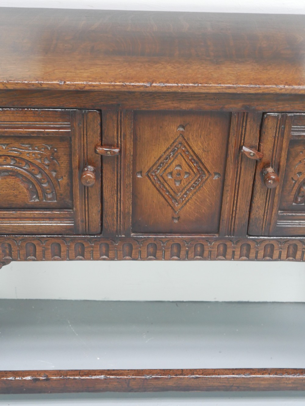 A small dark oak carved sideboard featuring panelled cupboard doors with carved arch motifs, iron hinges, and turned legs with a stretcher base. View III