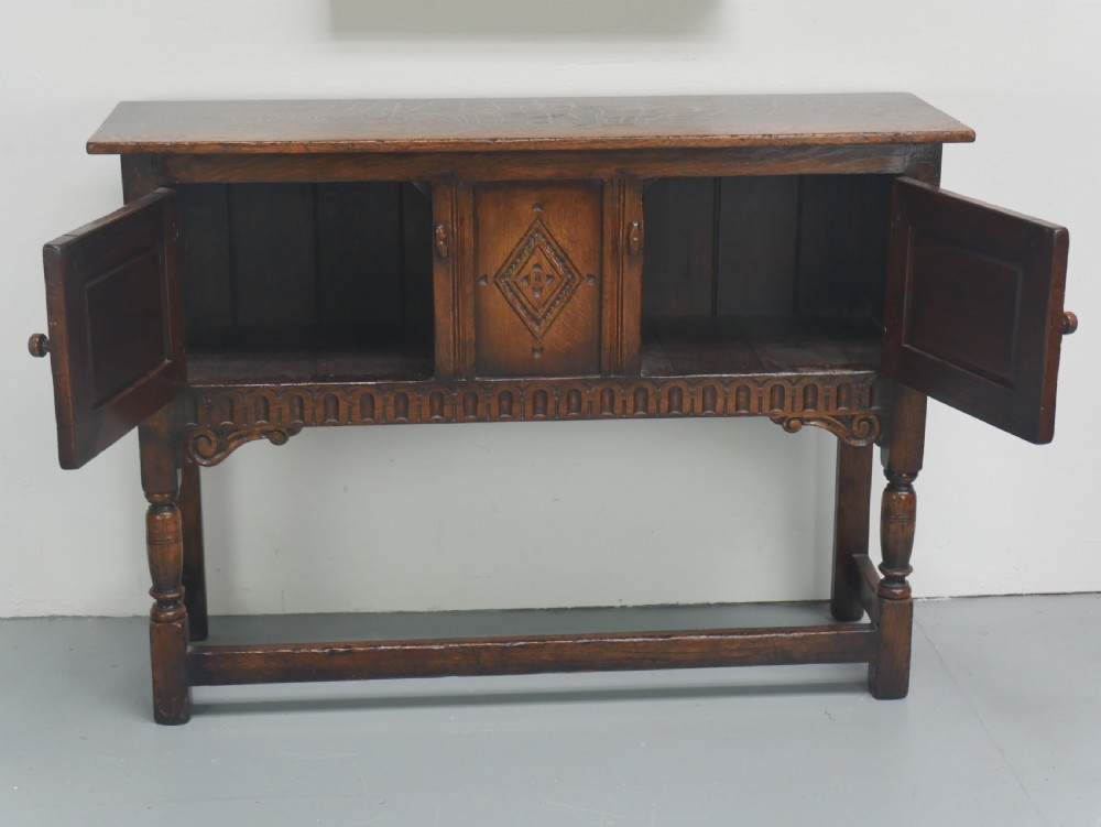 A small dark oak carved sideboard featuring panelled cupboard doors with carved arch motifs, iron hinges, and turned legs with a stretcher base. View IV