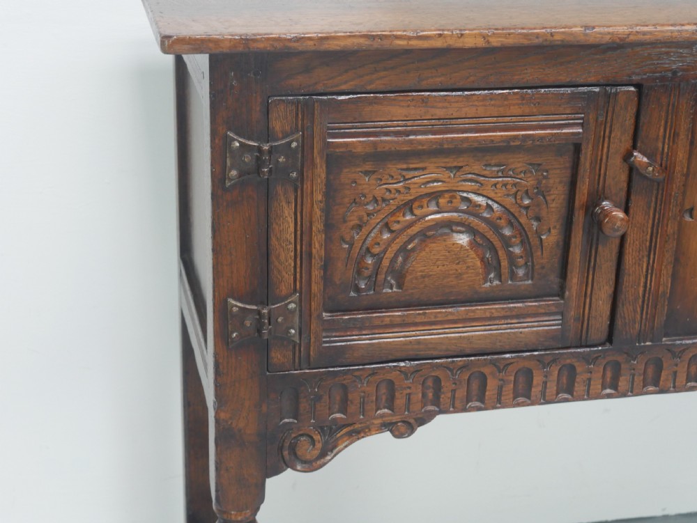 A small dark oak carved sideboard featuring panelled cupboard doors with carved arch motifs, iron hinges, and turned legs with a stretcher base. View V