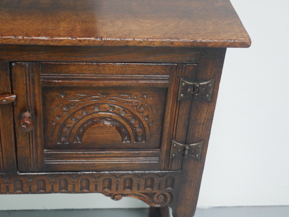 A small dark oak carved sideboard featuring panelled cupboard doors with carved arch motifs, iron hinges, and turned legs with a stretcher base. View VI