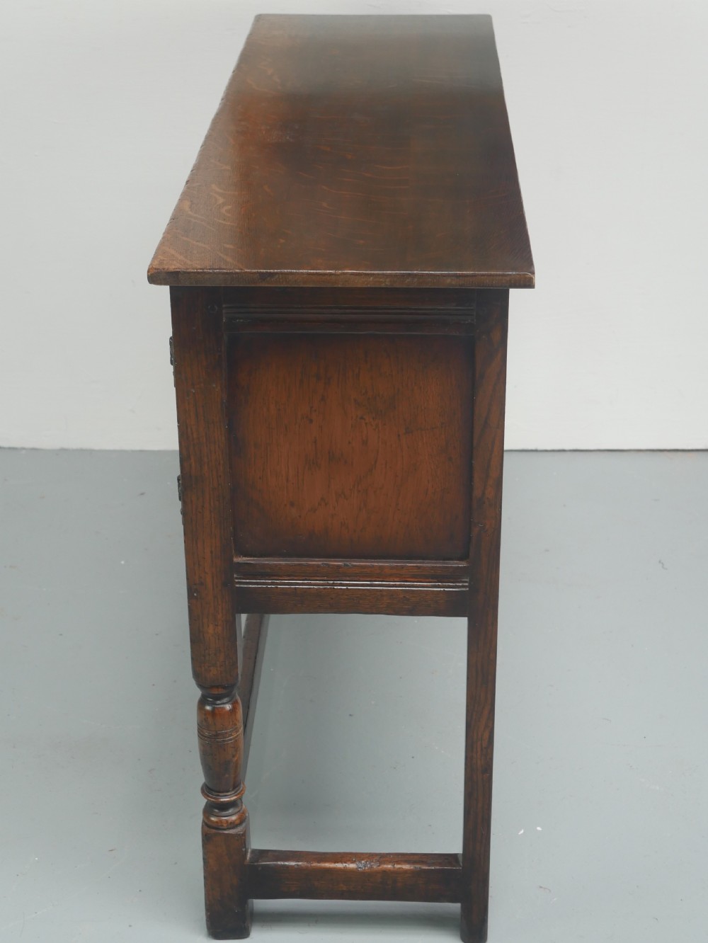 A small dark oak carved sideboard featuring panelled cupboard doors with carved arch motifs, iron hinges, and turned legs with a stretcher base. View VII