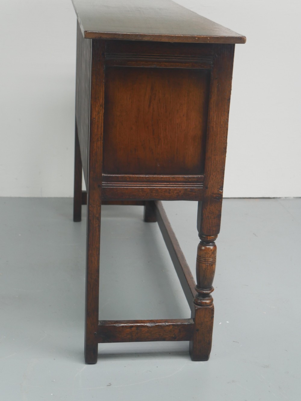 A small dark oak carved sideboard featuring panelled cupboard doors with carved arch motifs, iron hinges, and turned legs with a stretcher base. View X