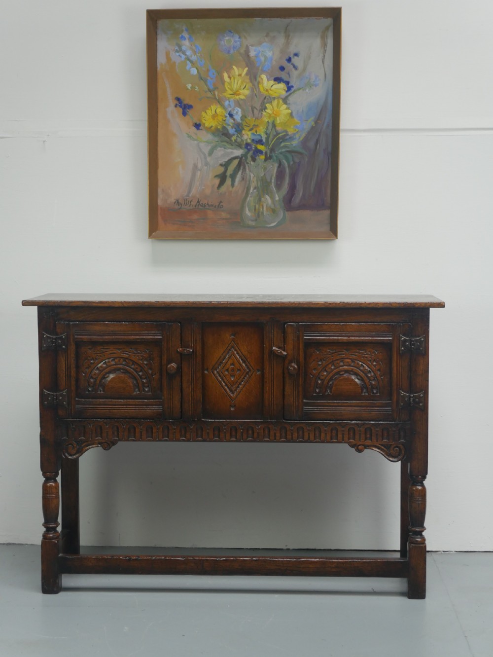 A small dark oak carved sideboard featuring panelled cupboard doors with carved arch motifs, iron hinges, and turned legs with a stretcher base. View XIII