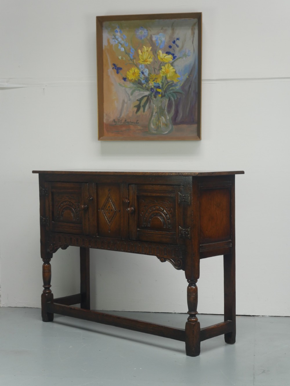A small dark oak carved sideboard featuring panelled cupboard doors with carved arch motifs, iron hinges, and turned legs with a stretcher base. View XIV