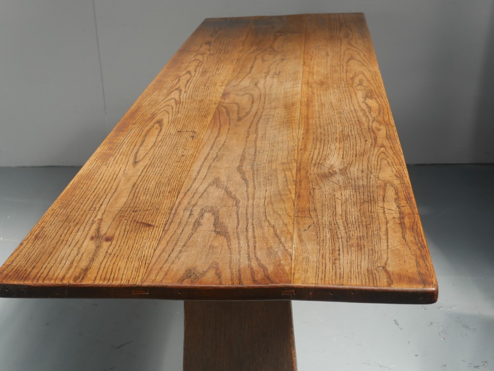 A long solid oak refectory dining table featuring a plank top, shaped trestle end supports, and a central pegged stretcher. View III