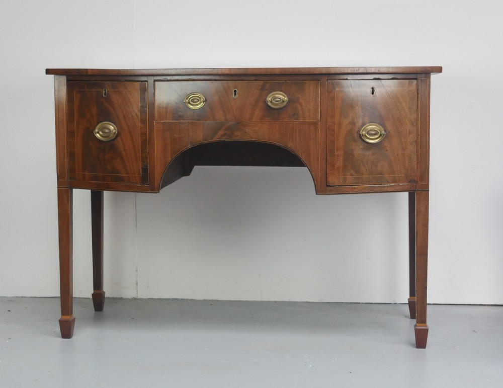 A small Georgian bowfront mahogany sideboard featuring an arched central apron, two side drawers with oval brass handles, and square tapered legs. View XV