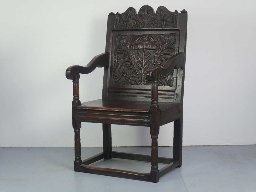 A dark oak 17th-century wainscot armchair featuring a relief-carved back panel, turned front legs, and flat armrests.