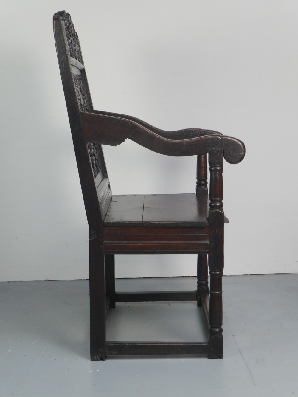 A dark oak 17th-century wainscot armchair featuring a relief-carved back panel, turned front legs, and flat armrests. View X