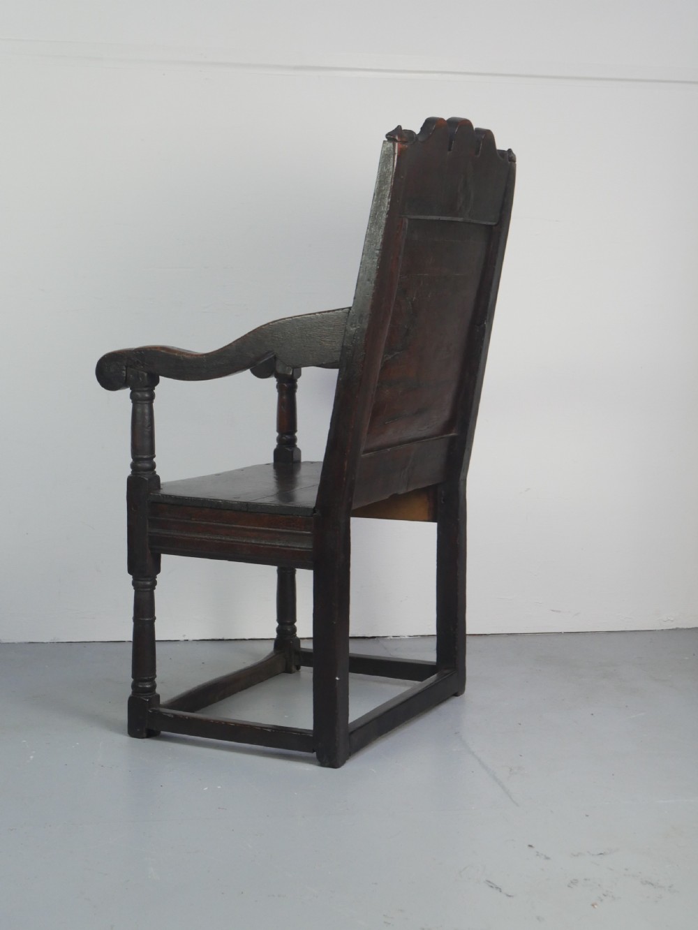 A dark oak 17th-century wainscot armchair featuring a relief-carved back panel, turned front legs, and flat armrests. View XII