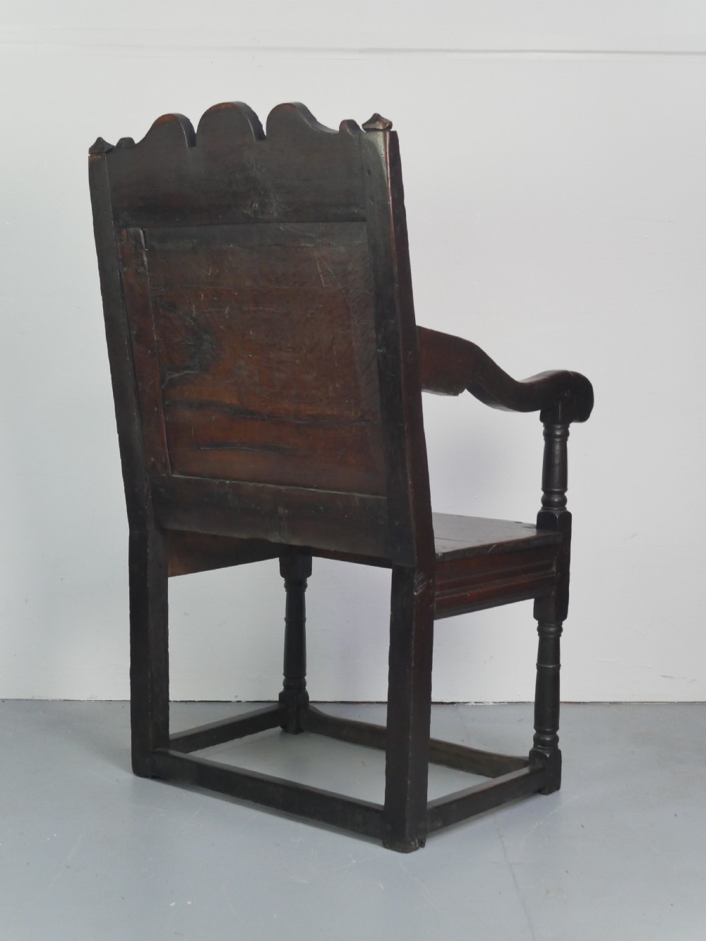 A dark oak 17th-century wainscot armchair featuring a relief-carved back panel, turned front legs, and flat armrests. View XV
