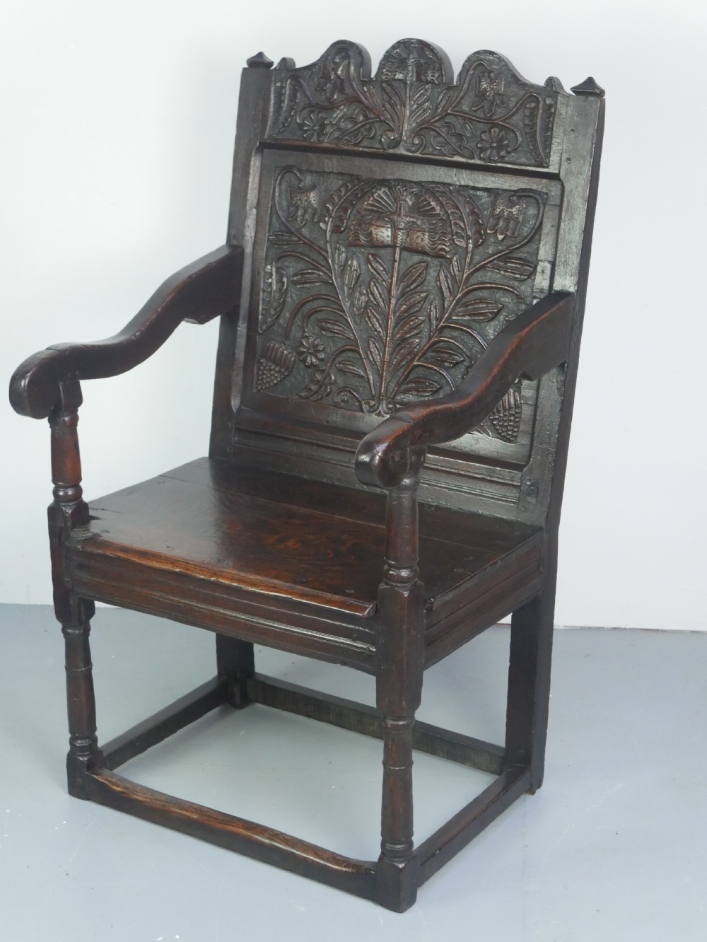 A dark oak 17th-century wainscot armchair featuring a relief-carved back panel, turned front legs, and flat armrests. View XXIII