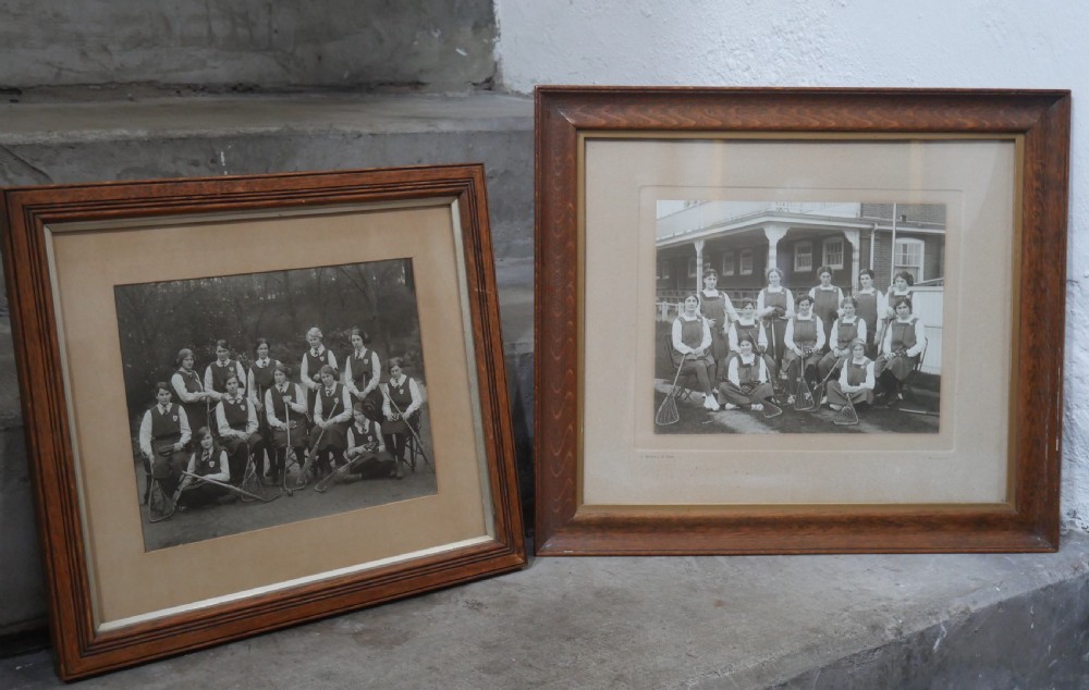 A framed monochrome photograph showing a 1914 womens lacrosse team seated with equipment, set within a moulded oak wood frame.