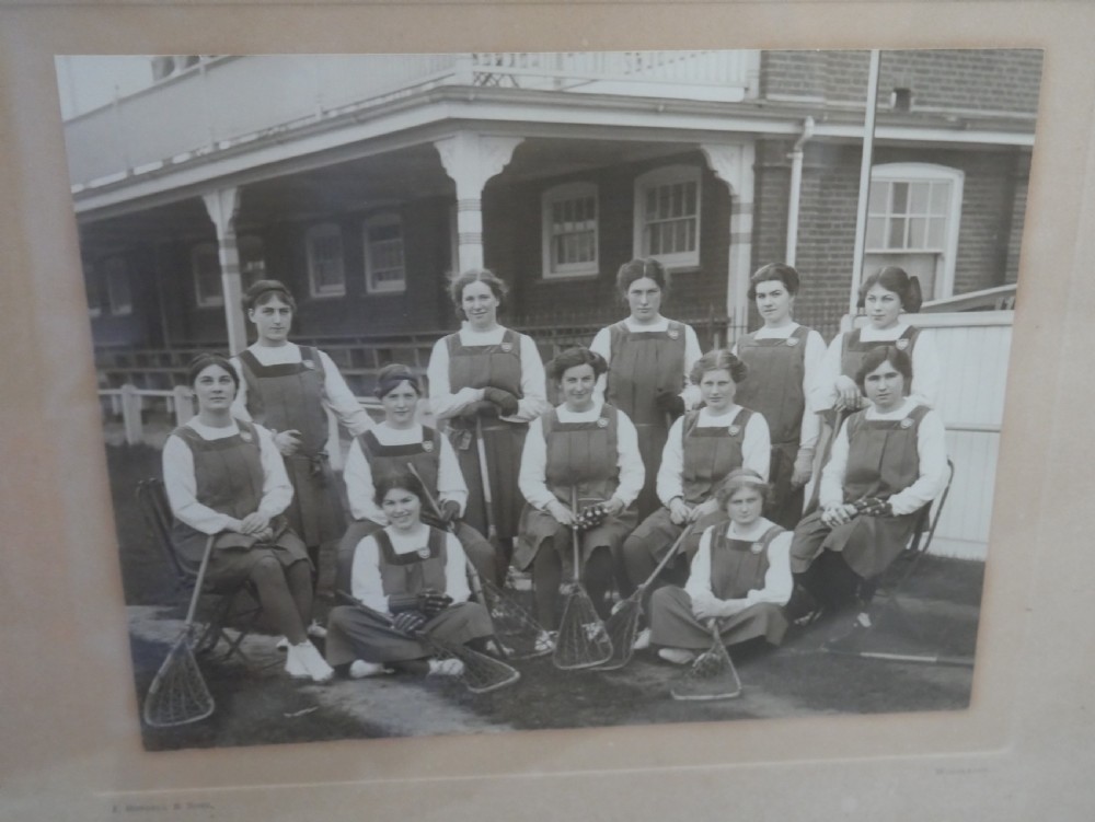 A framed monochrome photograph showing a 1914 womens lacrosse team seated with equipment, set within a moulded oak wood frame. View II
