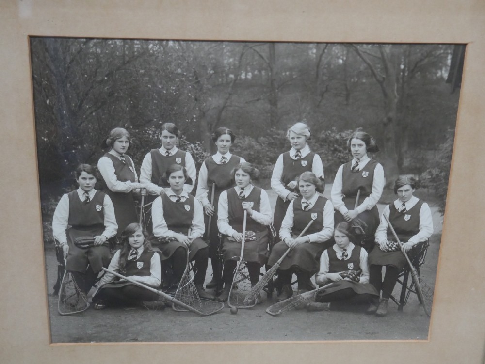 A framed monochrome photograph showing a 1914 womens lacrosse team seated with equipment, set within a moulded oak wood frame. View III