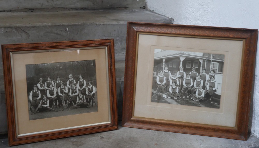 A framed monochrome photograph showing a 1914 womens lacrosse team seated with equipment, set within a moulded oak wood frame. View VI