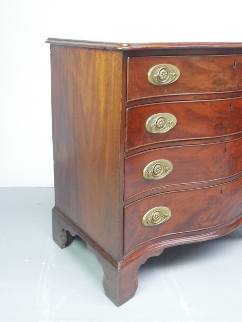 A Georgian mahogany chest of drawers featuring a wave-like serpentine front, four graduated drawers with oval brass handles, and bracket feet. View III