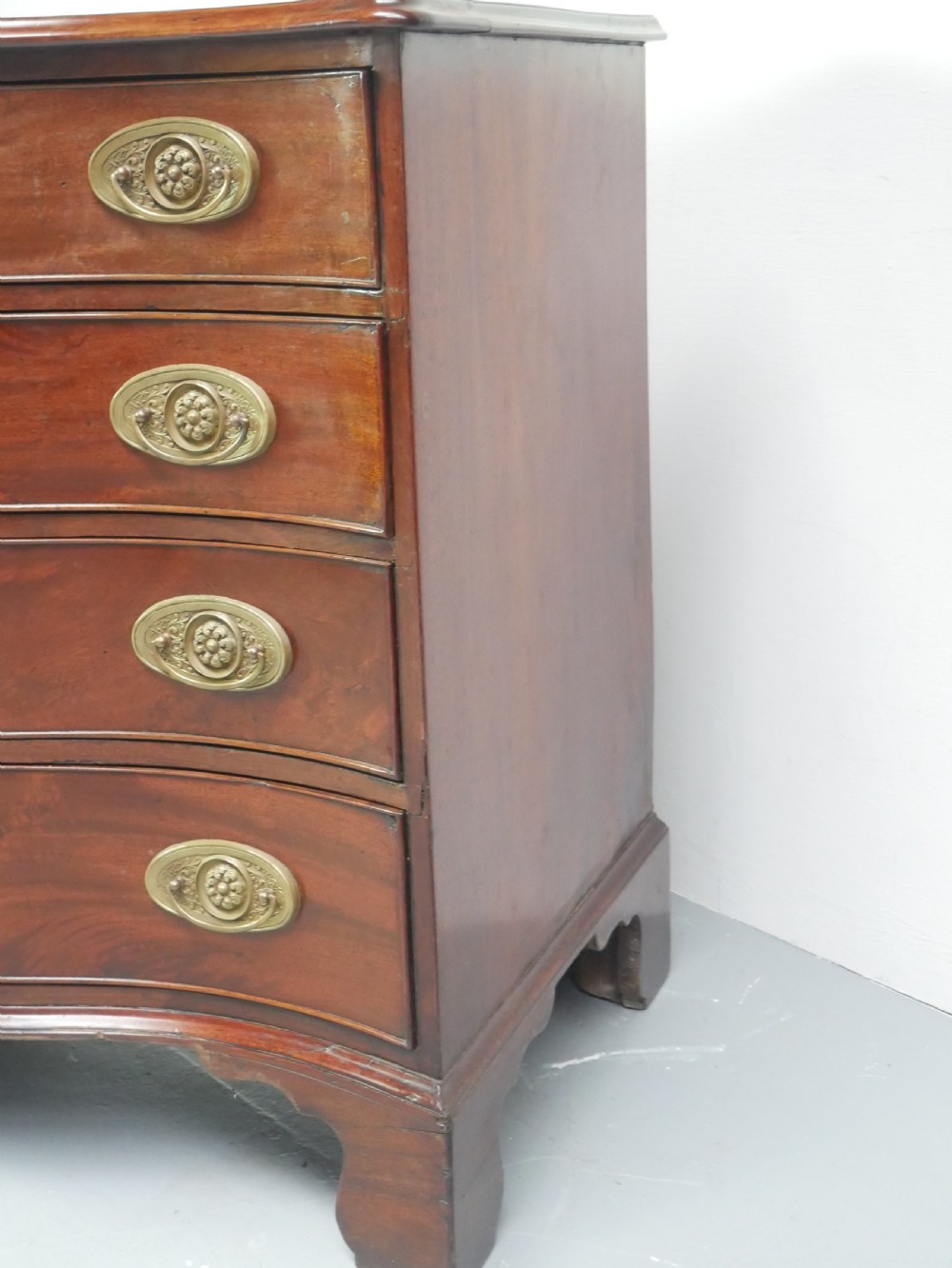 A Georgian mahogany chest of drawers featuring a wave-like serpentine front, four graduated drawers with oval brass handles, and bracket feet. View IV