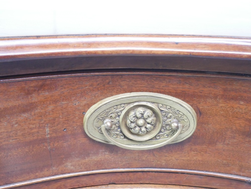 A Georgian mahogany chest of drawers featuring a wave-like serpentine front, four graduated drawers with oval brass handles, and bracket feet. View V