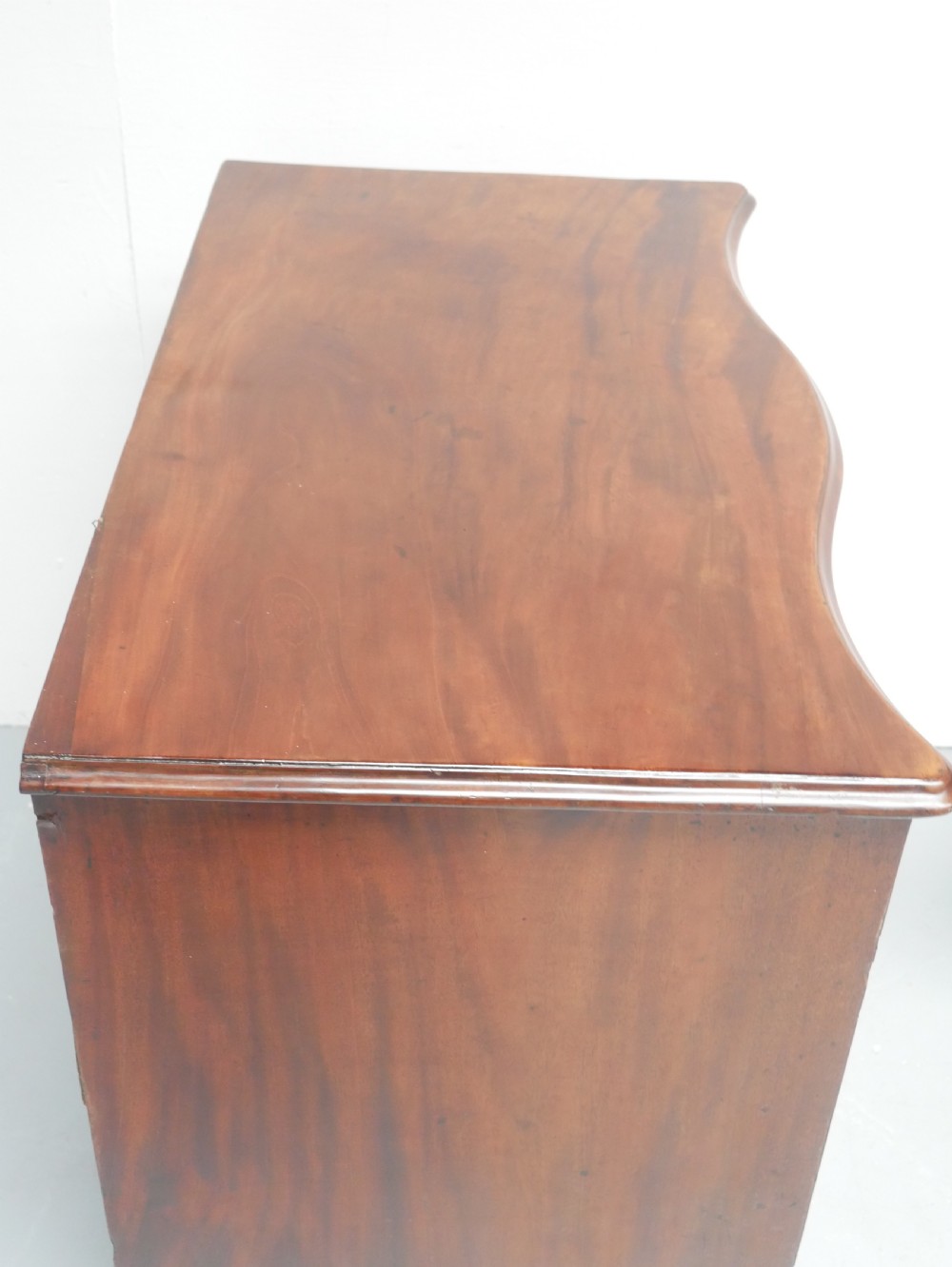 A Georgian mahogany chest of drawers featuring a wave-like serpentine front, four graduated drawers with oval brass handles, and bracket feet. View VI