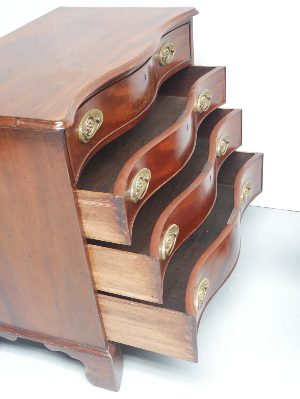 A Georgian mahogany chest of drawers featuring a wave-like serpentine front, four graduated drawers with oval brass handles, and bracket feet. View VIII
