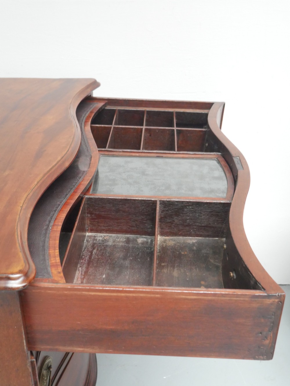 A Georgian mahogany chest of drawers featuring a wave-like serpentine front, four graduated drawers with oval brass handles, and bracket feet. View X