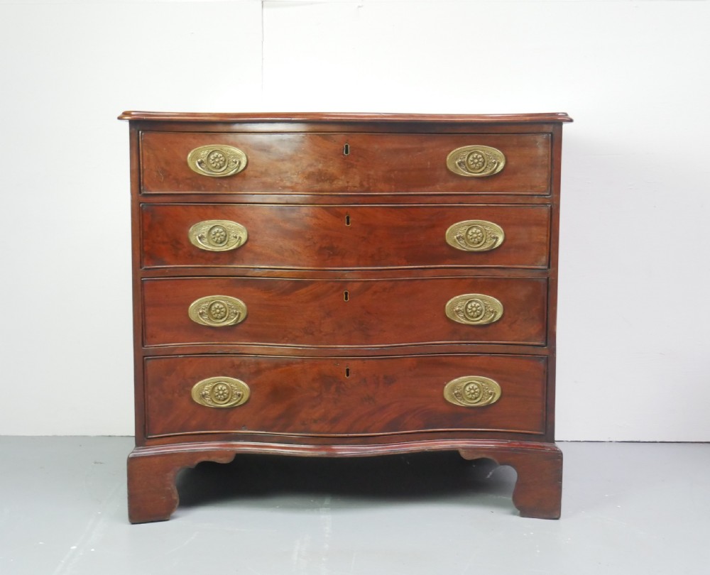 A Georgian mahogany chest of drawers featuring a wave-like serpentine front, four graduated drawers with oval brass handles, and bracket feet. View XIV