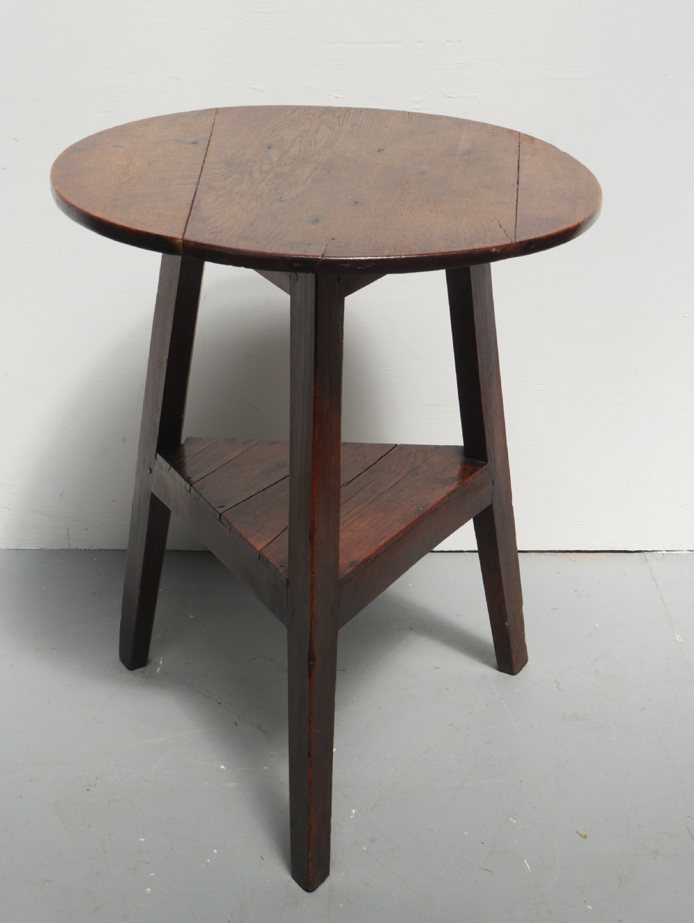 A dark Georgian oak cricket table featuring a circular plank top, three tapered legs, and a lower triangular shelf.