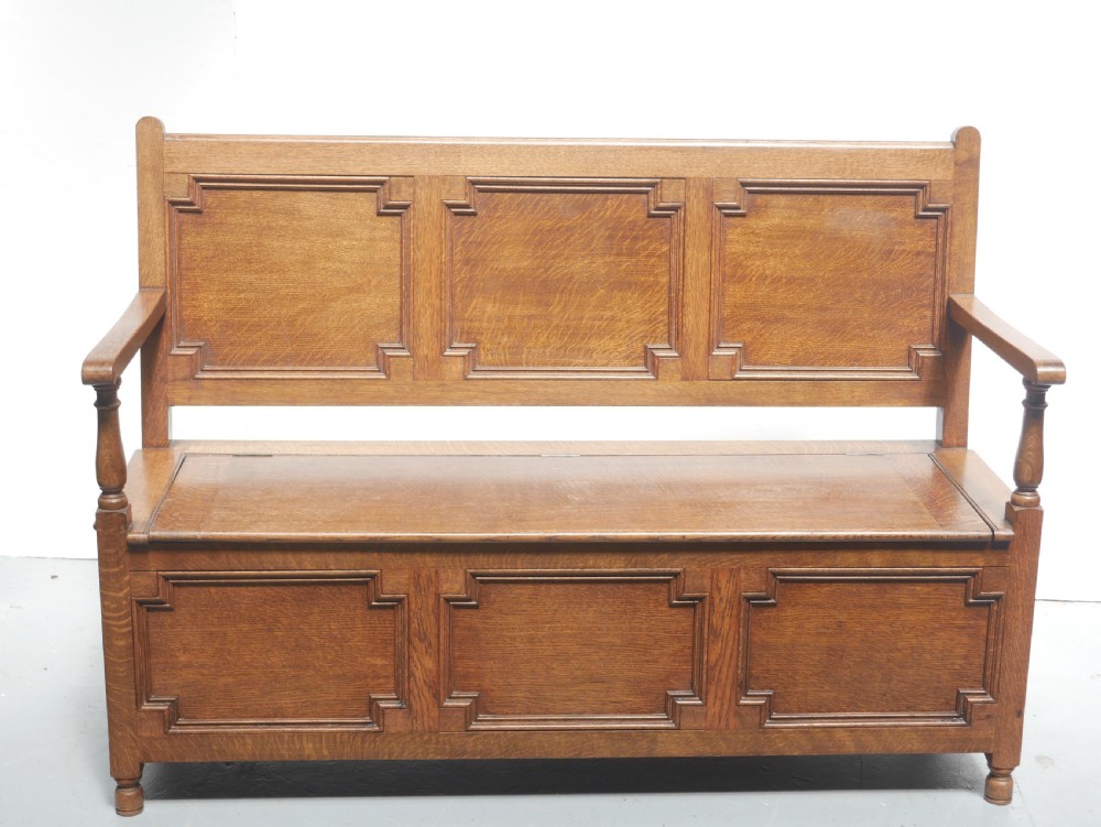 A medium brown oak hall bench featuring a tri-panel backrest, turned armrest supports, and a hinged lid chest base.