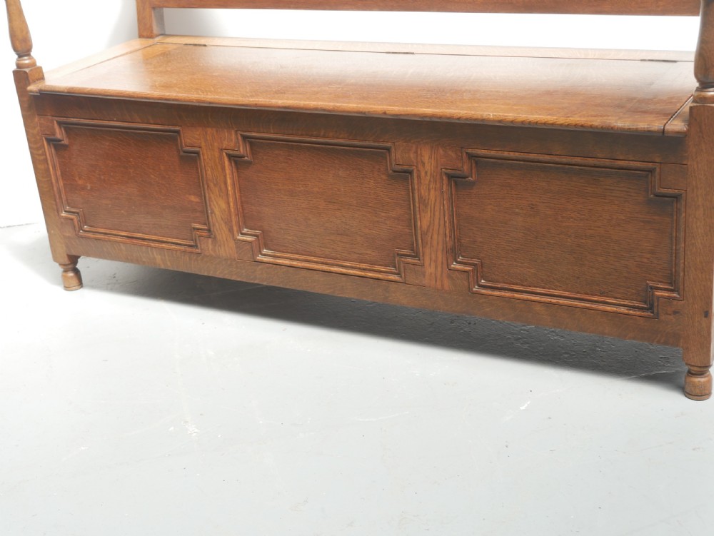 A medium brown oak hall bench featuring a tri-panel backrest, turned armrest supports, and a hinged lid chest base. View II