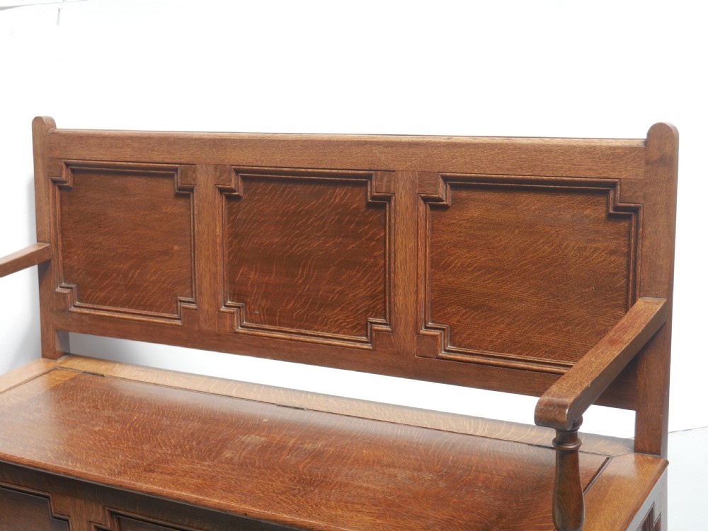 A medium brown oak hall bench featuring a tri-panel backrest, turned armrest supports, and a hinged lid chest base. View III