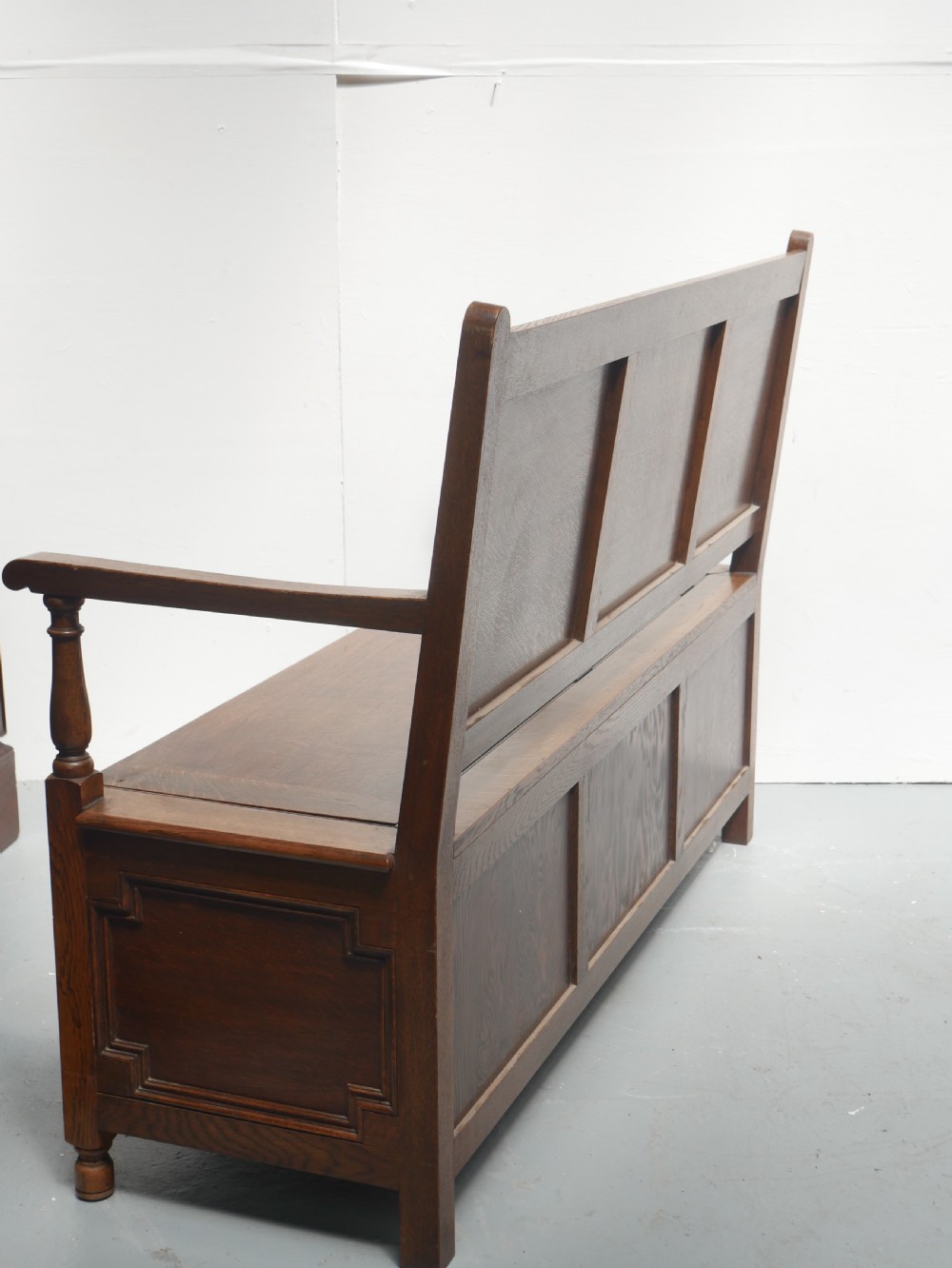A medium brown oak hall bench featuring a tri-panel backrest, turned armrest supports, and a hinged lid chest base. View VIII