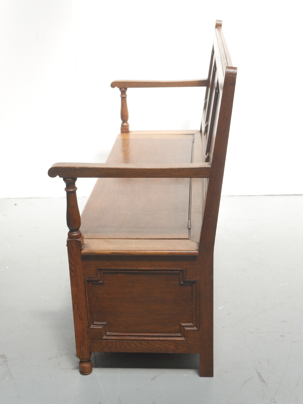 A medium brown oak hall bench featuring a tri-panel backrest, turned armrest supports, and a hinged lid chest base. View XI