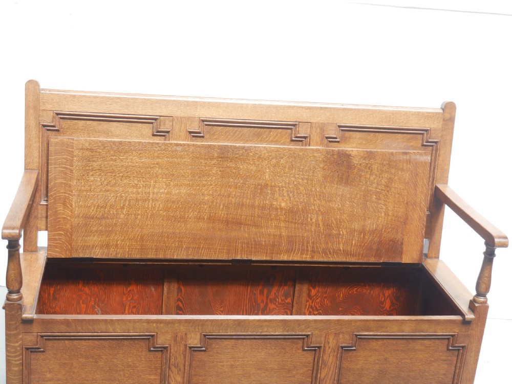 A medium brown oak hall bench featuring a tri-panel backrest, turned armrest supports, and a hinged lid chest base. View XII