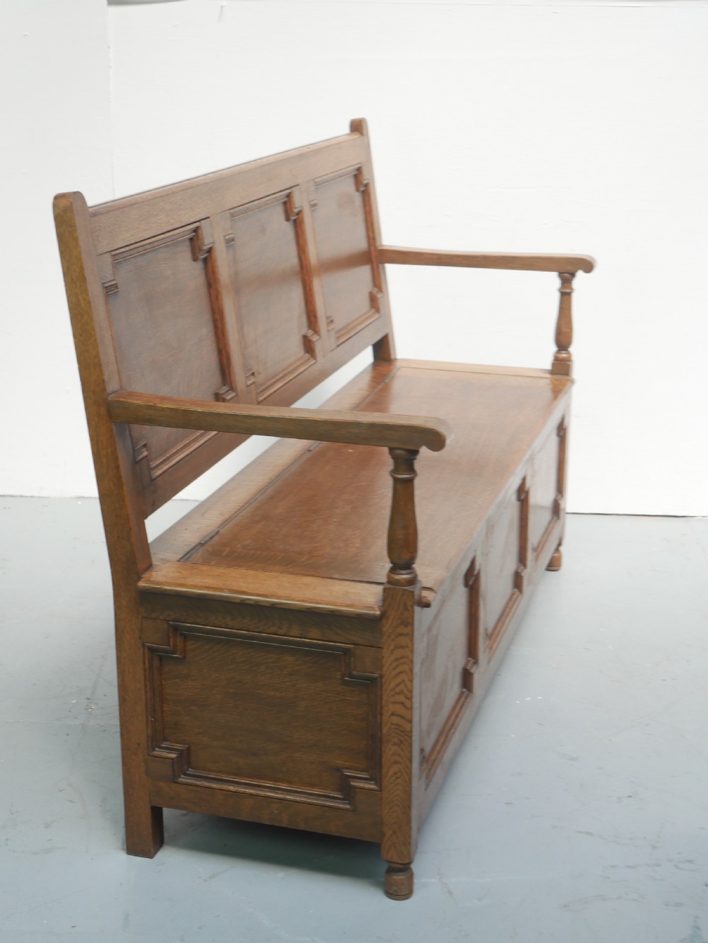 A medium brown oak hall bench featuring a tri-panel backrest, turned armrest supports, and a hinged lid chest base. View XIV